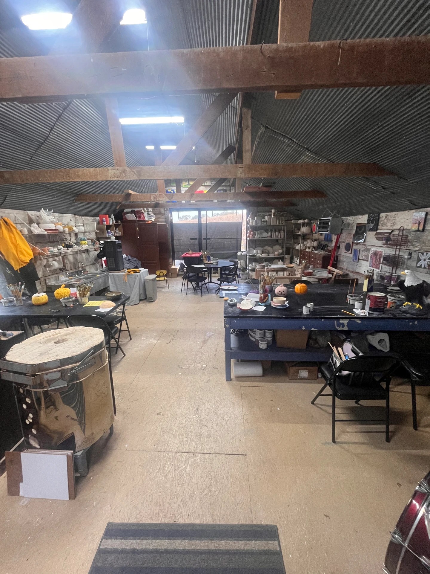 Inside Kask Studio in Denison, Iowa: a round kiln on a rolling base, shelves stacked with ceramic pieces waiting to be painted, and worktables set with paints, brushes, and clay